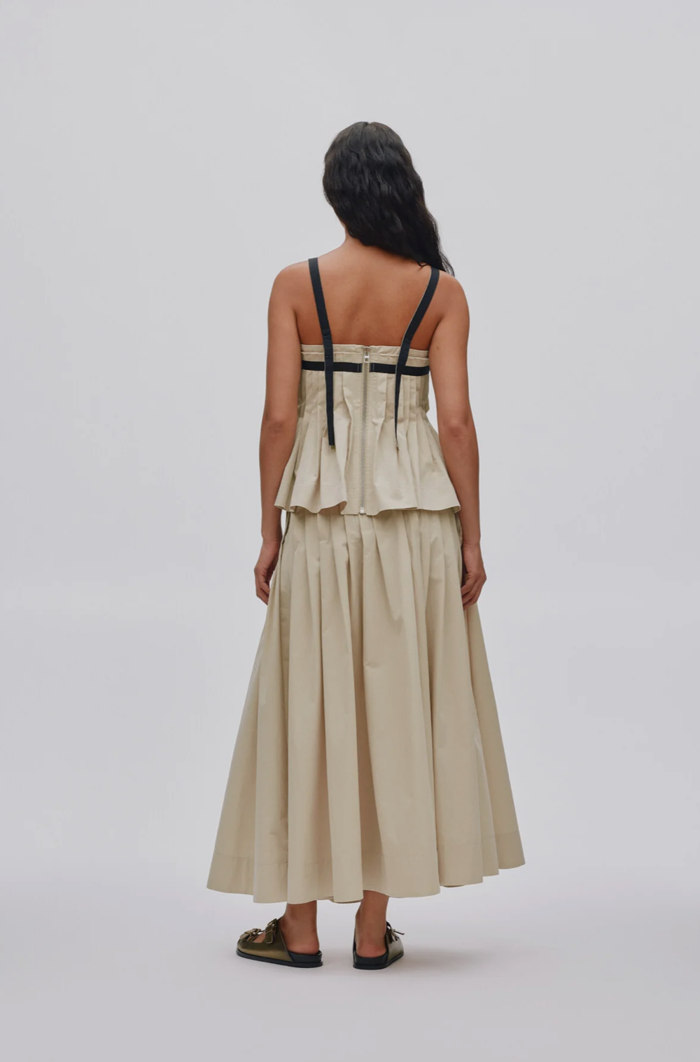 A woman viewed from the back wearing a sleeveless beige maxi dress with black shoulder straps and a ruffled bodice, paired with dark sandals, against a light gray studio background.
