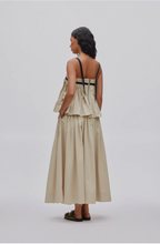 A woman shown from the back wearing a beige sleeveless maxi dress with thin black shoulder straps and a ruffled peplum waist, photographed against a light gray studio background.