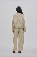 A model viewed from the back wearing a beige cropped jacket with a unique triangular back detail, matching wide-leg trousers, and black sandals, set against a plain light gray background.