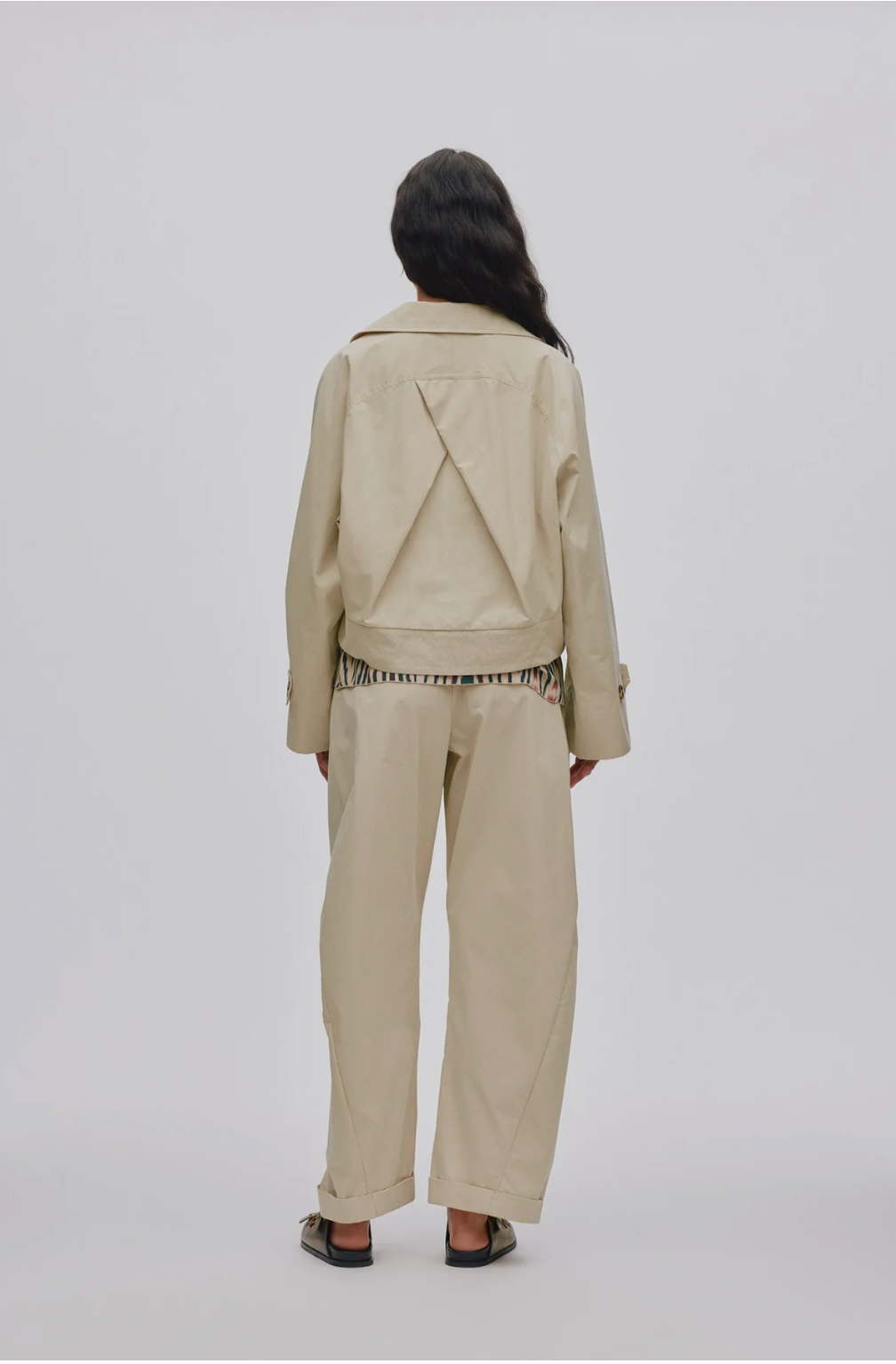 A model viewed from the back wearing a beige cropped jacket with a unique triangular back detail, matching wide-leg trousers, and black sandals, set against a plain light gray background.