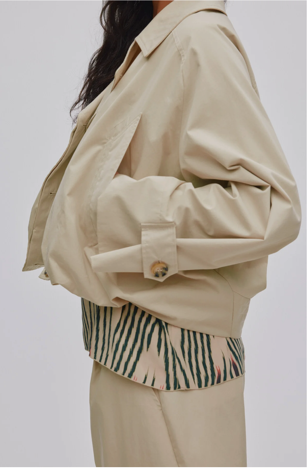 A person wearing an oversized light beige jacket over a cream and green striped shirt, paired with matching beige bottoms, photographed against a plain light gray studio background.