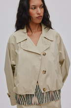 A woman wearing a light beige cropped utility jacket with gold buttons, layered over a green and beige striped top, against a plain light gray background.