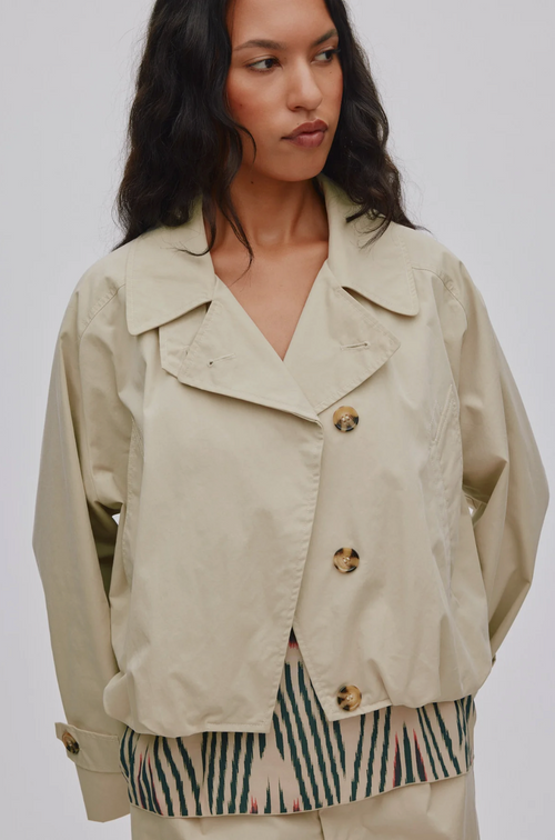 A woman wearing a light beige cropped utility jacket with gold buttons, layered over a green and beige striped top, against a plain light gray background.