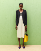 A woman standing against a green background, wearing a black slim-fit twill jacket with a V-neck and long sleeves, and a white dress with a top accessory. She is also holding a yellow bag and wearing purple shoes.