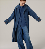 A woman modeling a long blue denim trench coat with a pointed collar, raglan sleeves, and cuff tabs, featuring a detached yoke at the back and welt pockets. She is also wearing blue denim pants.