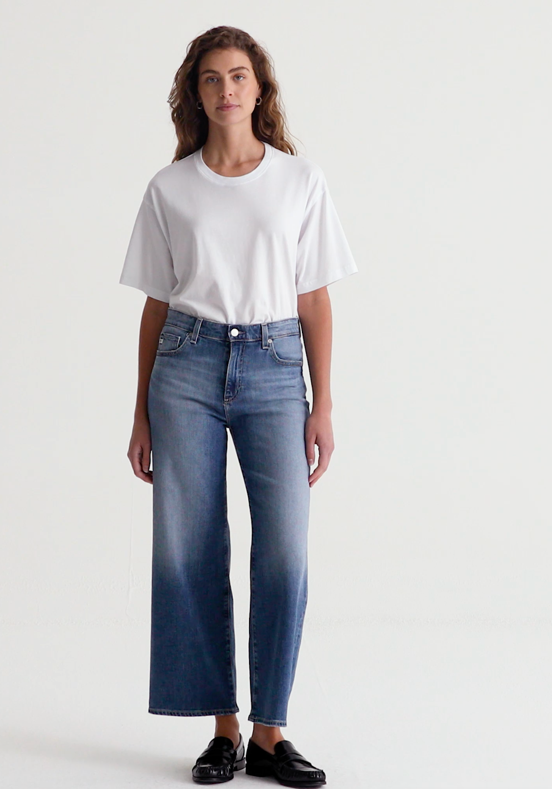 A person standing wearing mid-rise, wide-leg crop jeans with fraying hems and a light indigo wash. Also visible is a white t-shirt and black shoes.