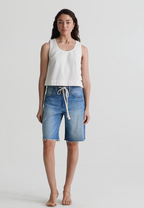 Woman wearing a white sleeveless top and blue denim shorts on a plain background