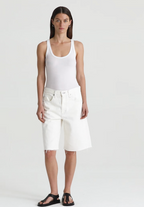 Woman wearing a white tank top and white ag jeans denim shorts on a plain background