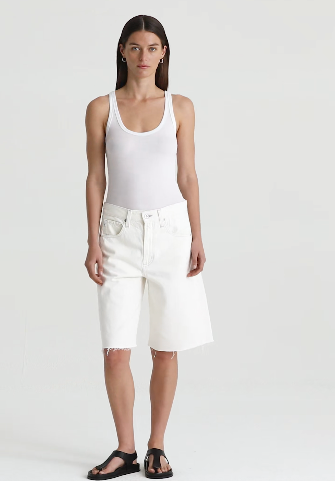 Woman wearing a white tank top and white ag jeans denim shorts on a plain background