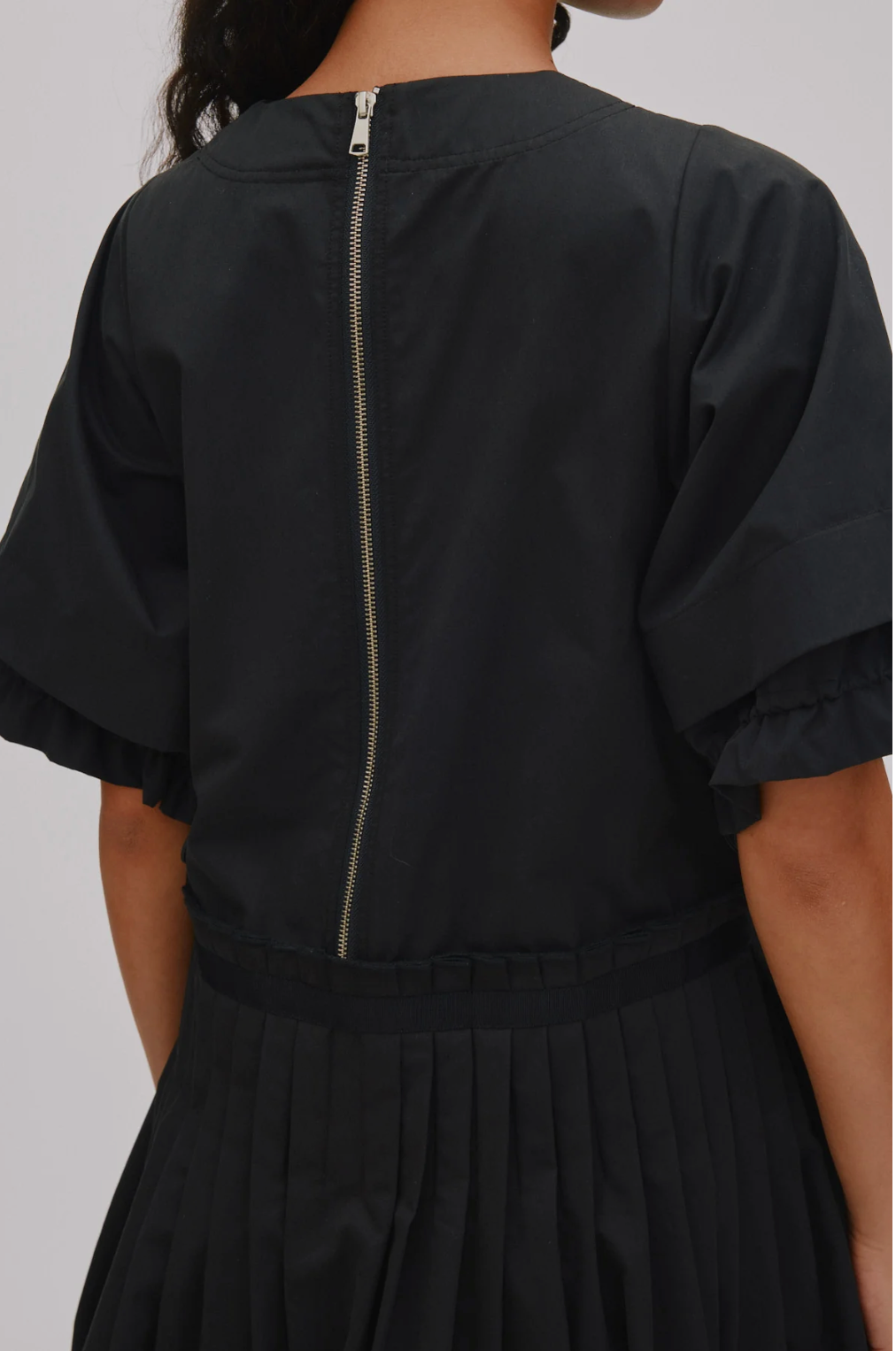 Close-up back view of a black dress with short flutter sleeves, a gathered waist, and a central zipper, shown on a model against a light gray background.