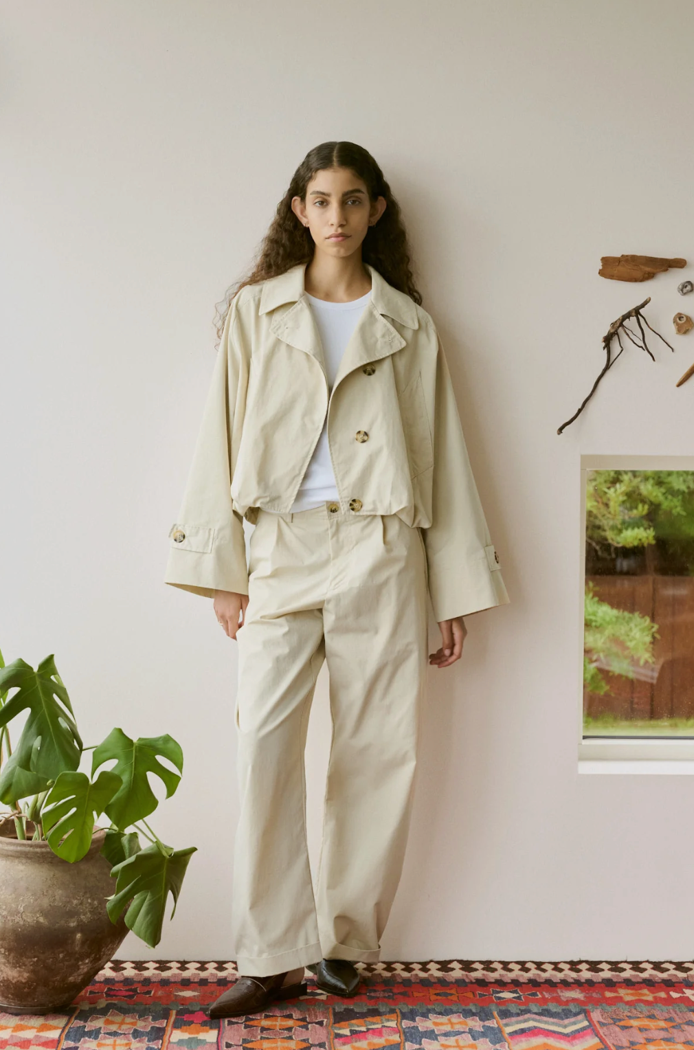A woman stands indoors wearing a light beige cropped jacket and matching wide-leg pants over a white top, with dark brown shoes. The room has a pale wall, a potted plant, a patterned rug, and small wall decorations.