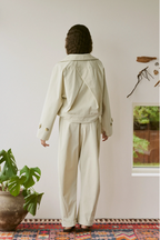 Person wearing a beige outfit standing in a room with a plant and window.