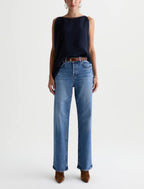 A model wearing a sleeveless dark navy top, blue wide-leg jeans, and brown suede boots, standing against a plain light gray studio background.