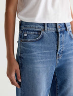 Close-up of a person wearing medium-wash blue denim jeans with a white top, showing the front pocket and waistband area.
