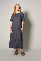 A woman modeling a loose-fitting, short-sleeved linen maxi dress in a dark blue-gray color, paired with dark sandals, against a plain light gray studio background.