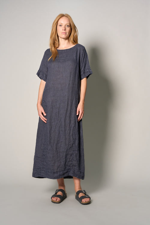 A woman modeling a loose-fitting, short-sleeved linen maxi dress in a dark blue-gray color, paired with dark sandals, against a plain light gray studio background.