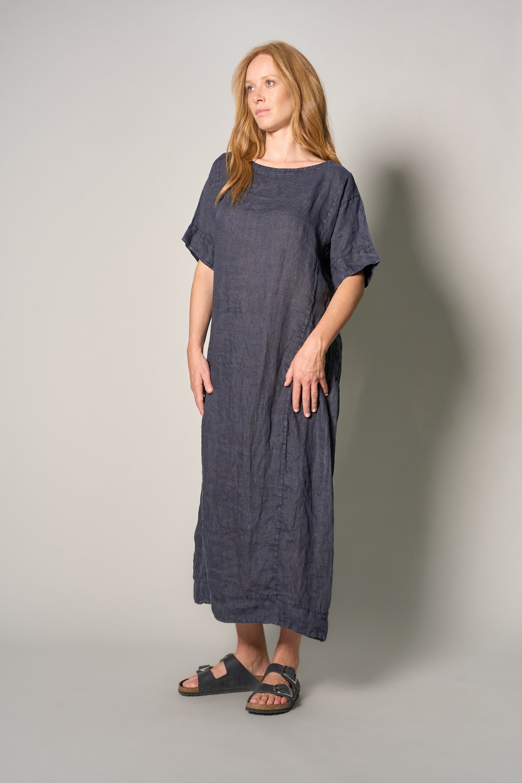 A woman modeling a loose-fitting, short-sleeved linen maxi dress in a dark blue-gray color, paired with black sandals, against a plain light gray studio background.