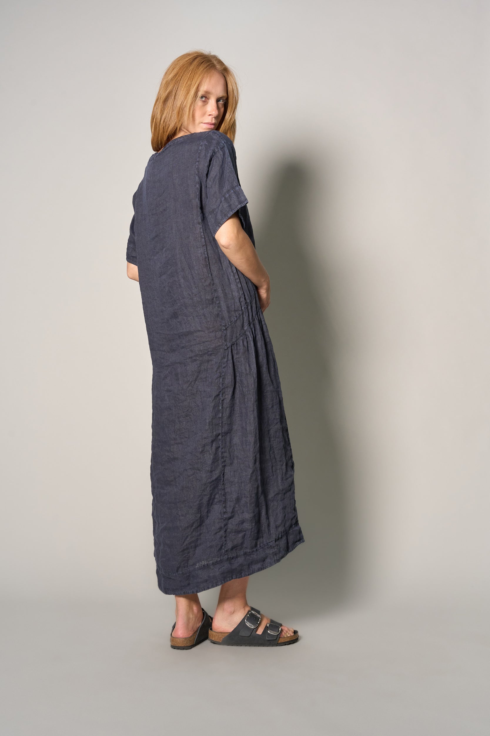 A woman with strawberry-blonde hair stands in profile wearing a loose, short-sleeved linen maxi dress in a dark navy blue color, paired with dark sandals, against a plain light gray studio background.