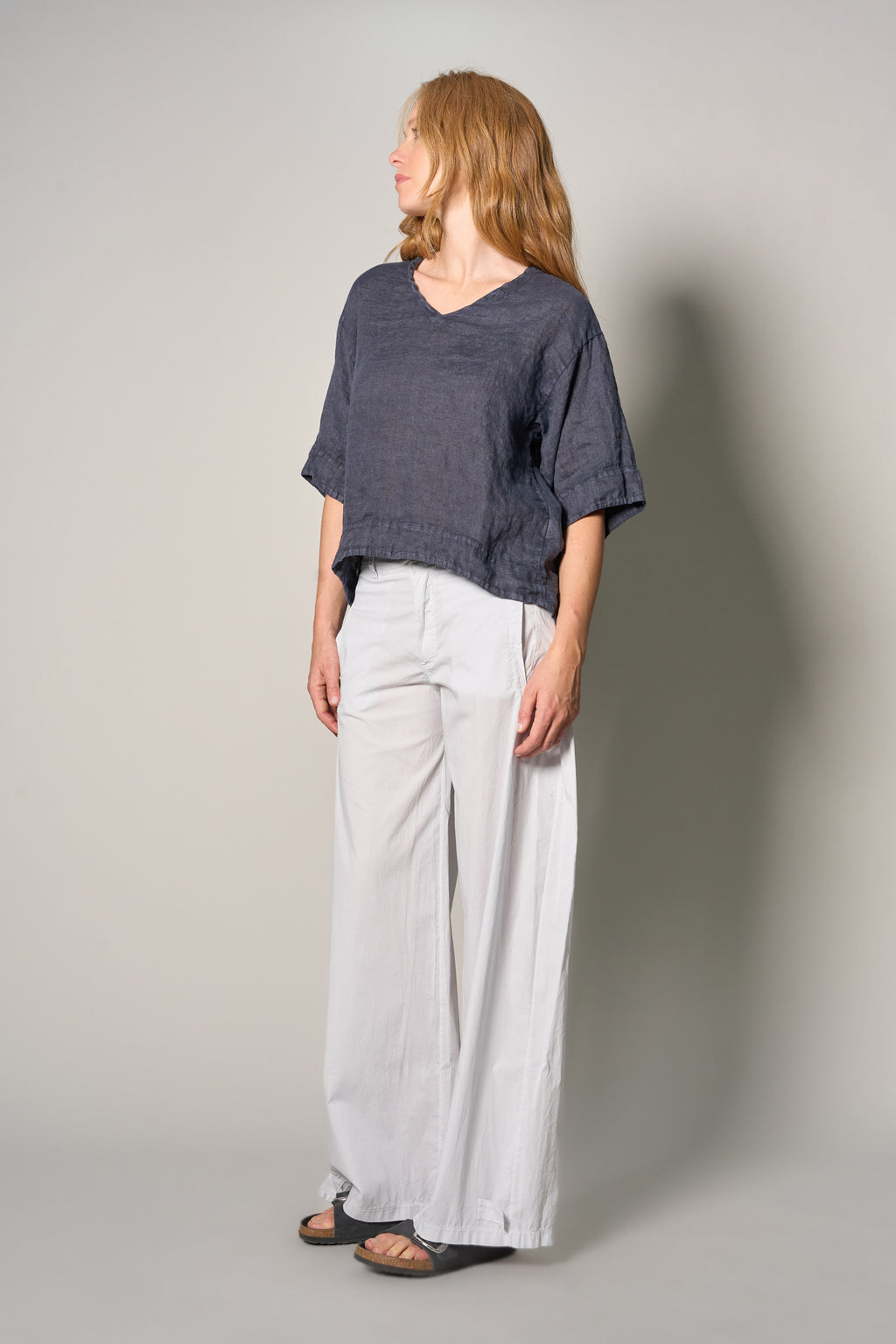 A woman stands in profile wearing a loose short-sleeve dark blue top and wide-leg light gray pants against a plain light gray studio background.