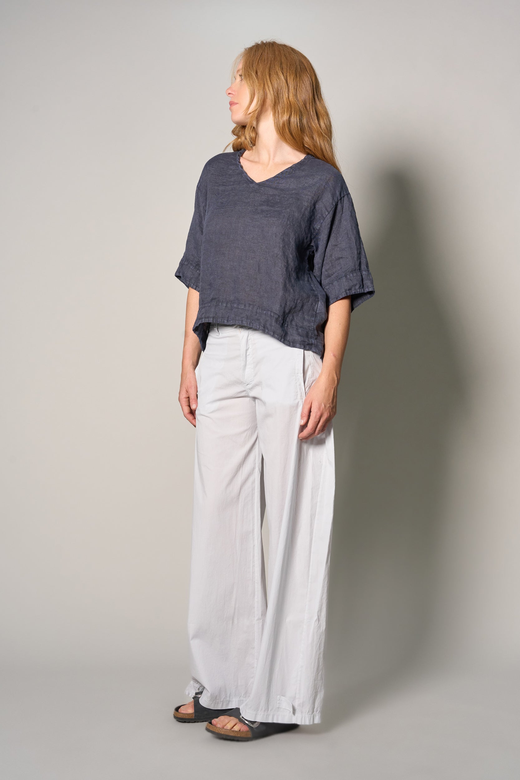 A woman stands in profile wearing a loose short-sleeve dark blue top and wide-leg light gray pants against a plain light gray studio background.