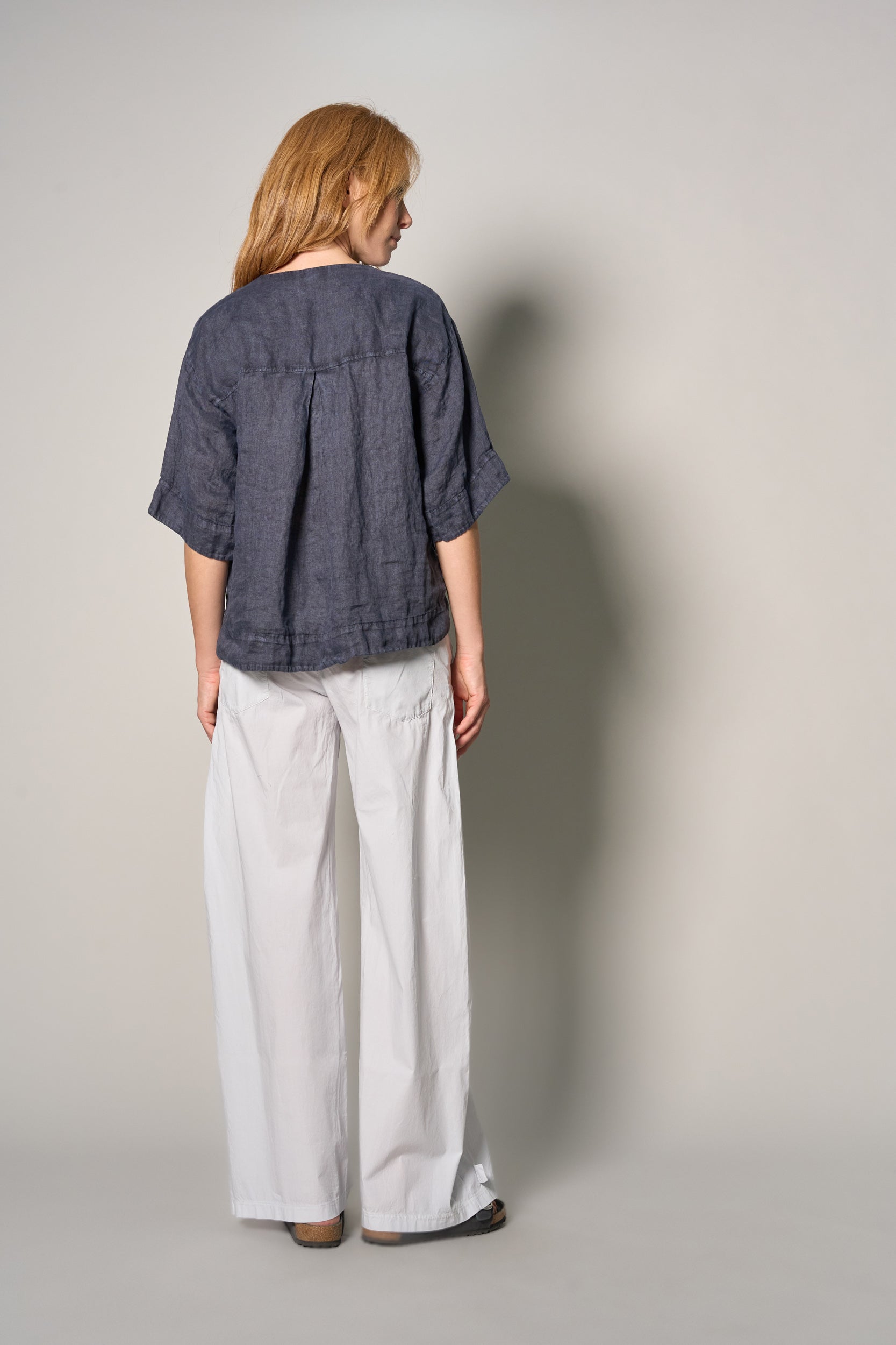 A woman is shown from the back wearing a dark slate blue linen short-sleeve top and white wide-leg trousers, standing against a neutral gray studio background.