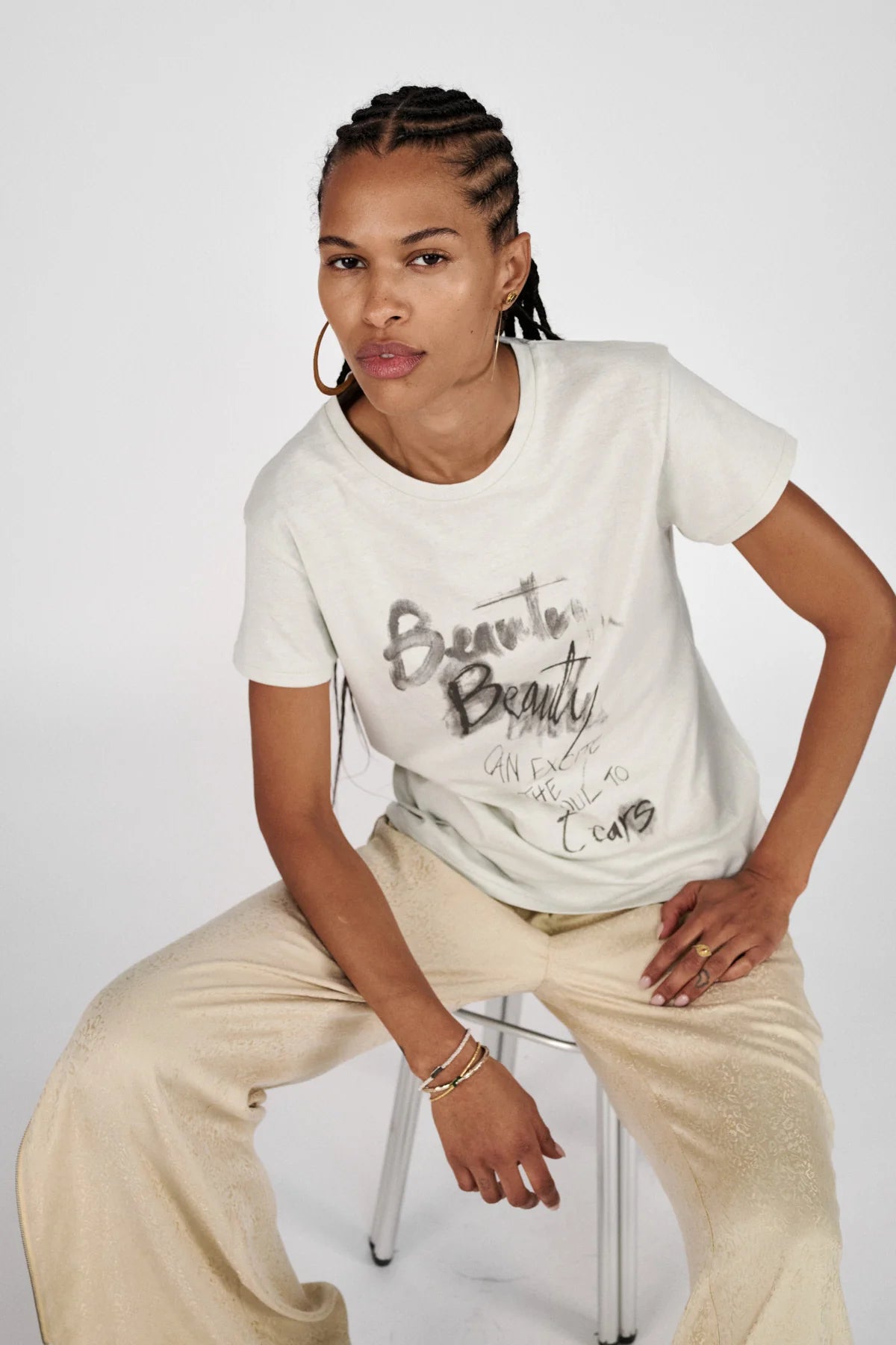 Woman wearing a white t-shirt with text and beige pants sitting on a stool against a white background