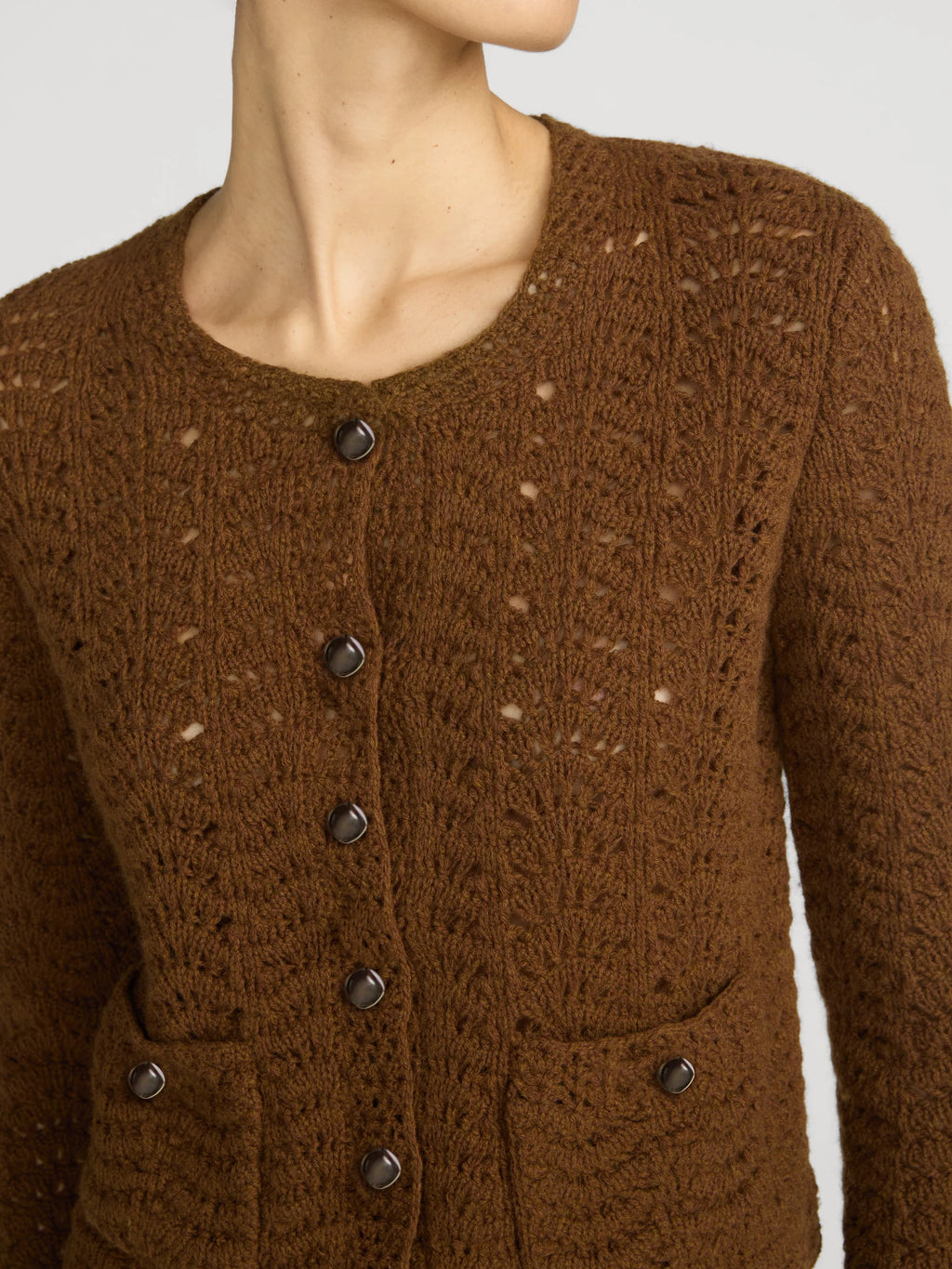 A close-up of a model wearing a brown textured knit cardigan with dark buttons and front pockets, shown against a light gray background.