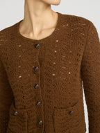 A close-up of a model wearing a brown textured knit cardigan with dark buttons and front pockets, shown against a light gray background.