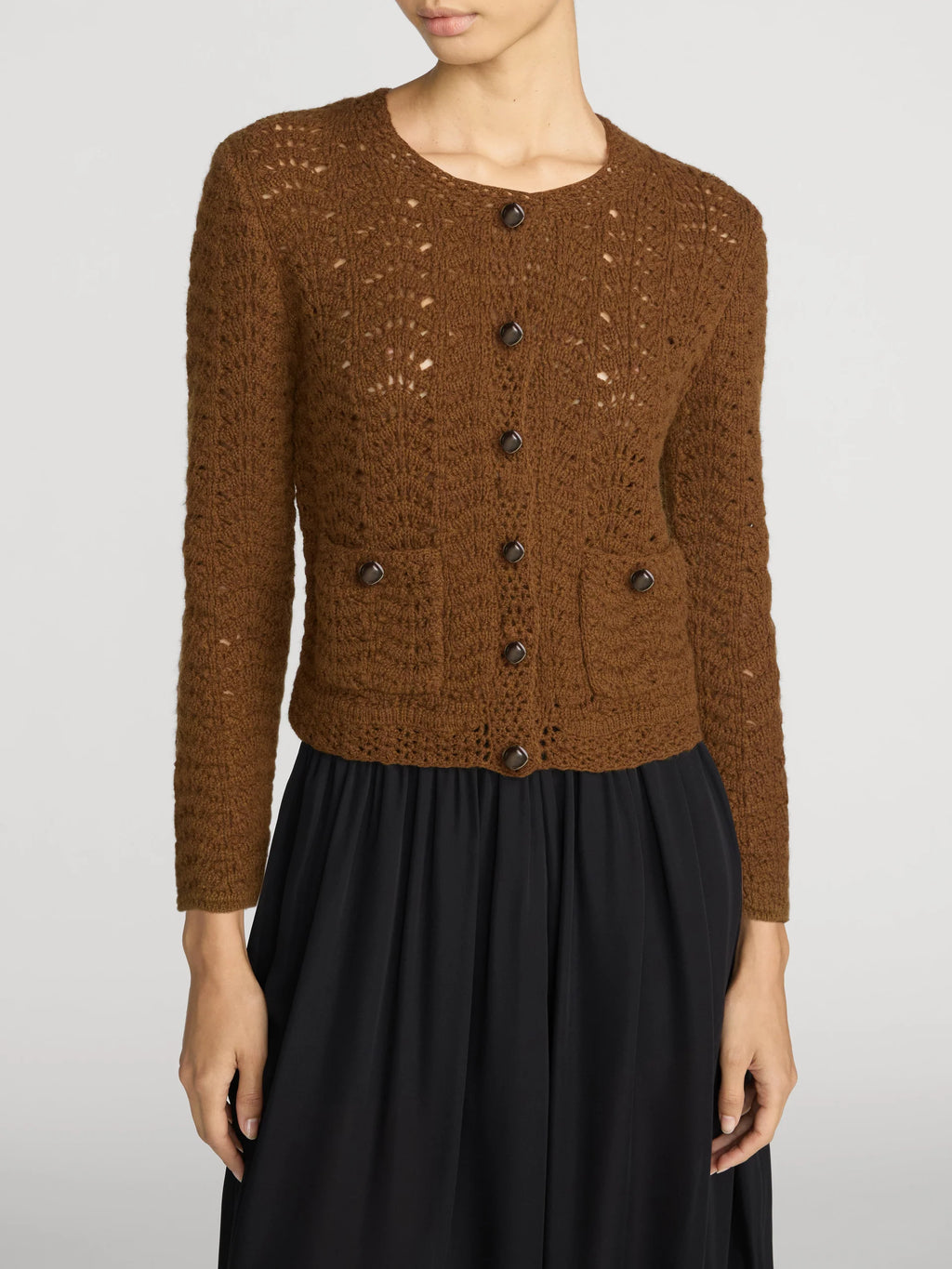 A woman wearing a brown open-knit button-front cardigan with dark buttons and a black skirt, photographed against a light gray studio background.