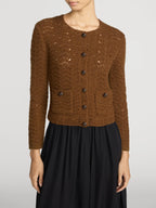 A woman wearing a brown open-knit button-front cardigan with dark buttons and a black skirt, photographed against a light gray studio background.