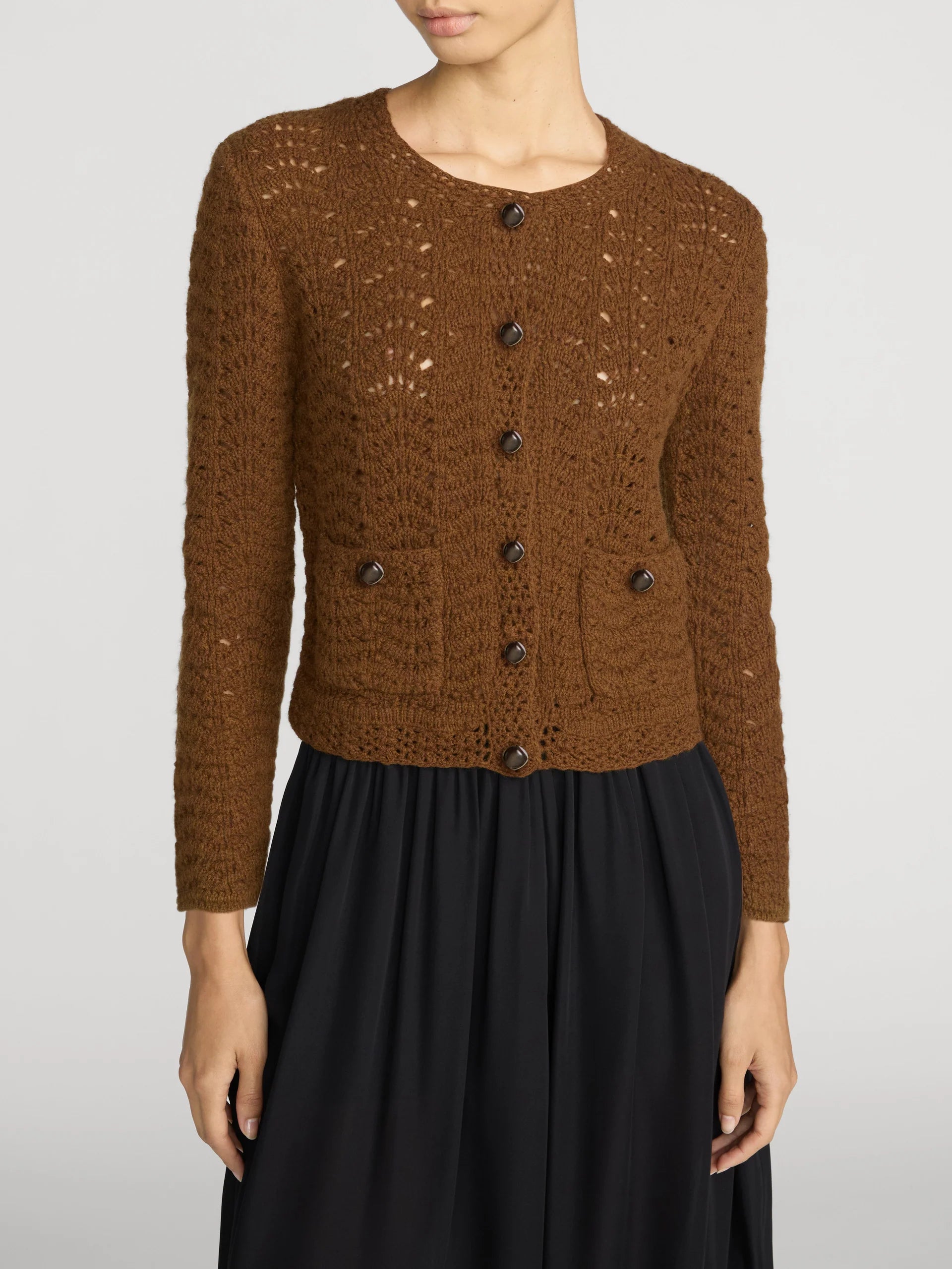 A woman wearing a brown open-knit button-front cardigan with dark buttons and a black skirt, photographed against a light gray studio background.