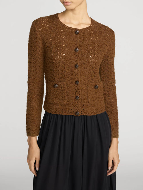 A woman wearing a brown open-knit button-front cardigan with dark buttons and a black skirt, photographed against a light gray studio background.