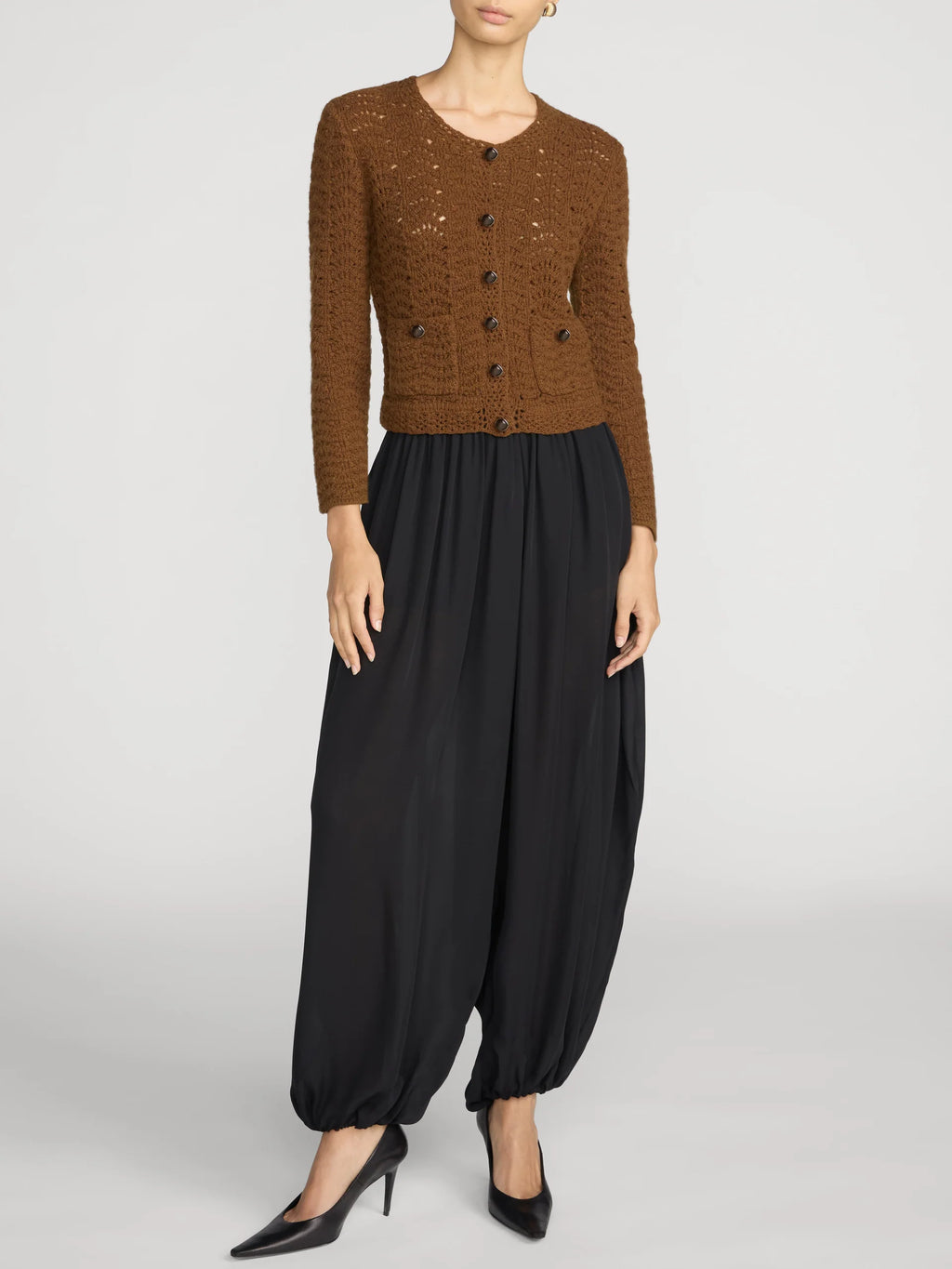 A model wearing a brown textured knit cardigan with dark buttons, paired with black balloon pants and black pointed-toe heels, photographed against a light gray studio background.