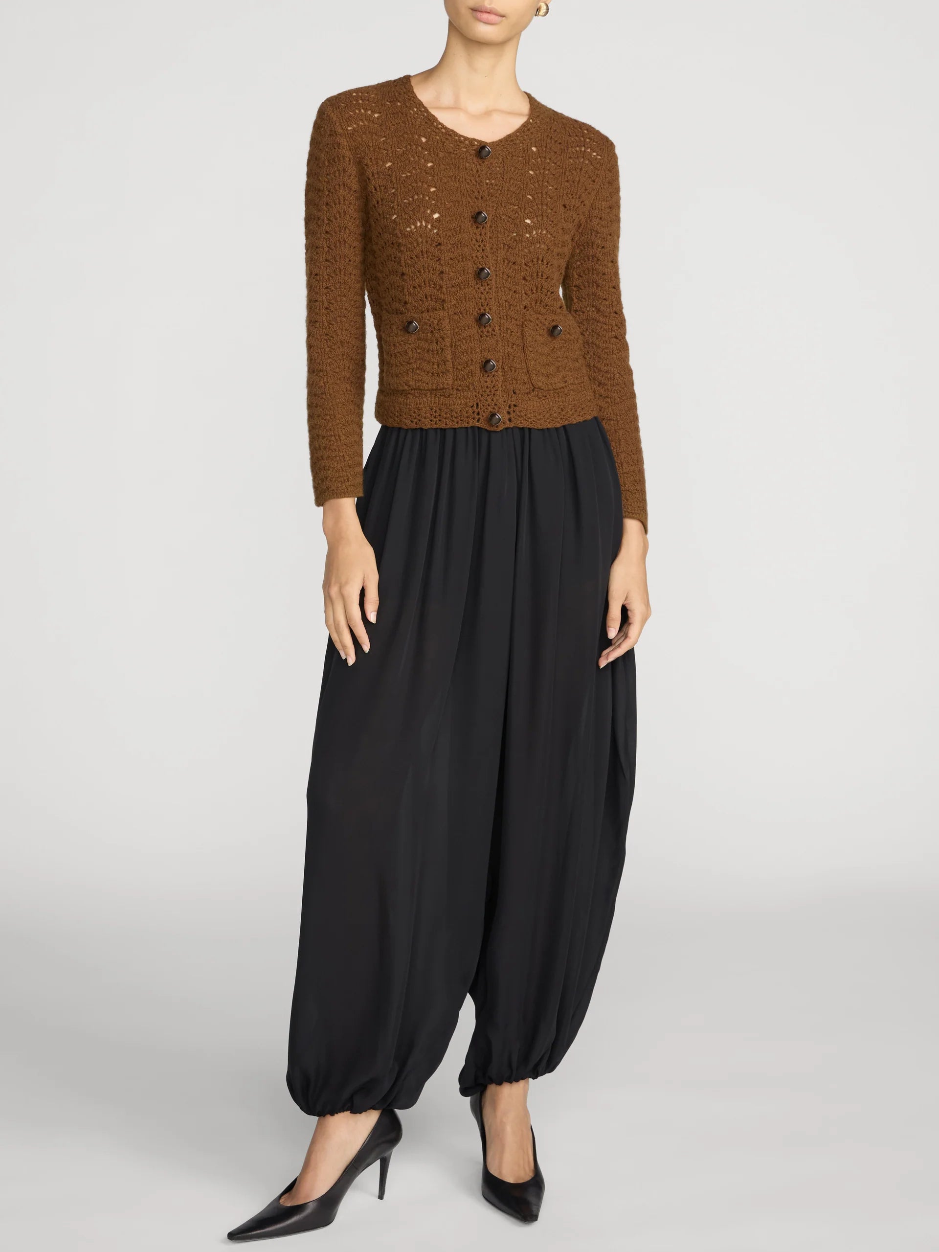A model wearing a brown textured knit cardigan with dark buttons, paired with black balloon pants and black pointed-toe heels, photographed against a light gray studio background.