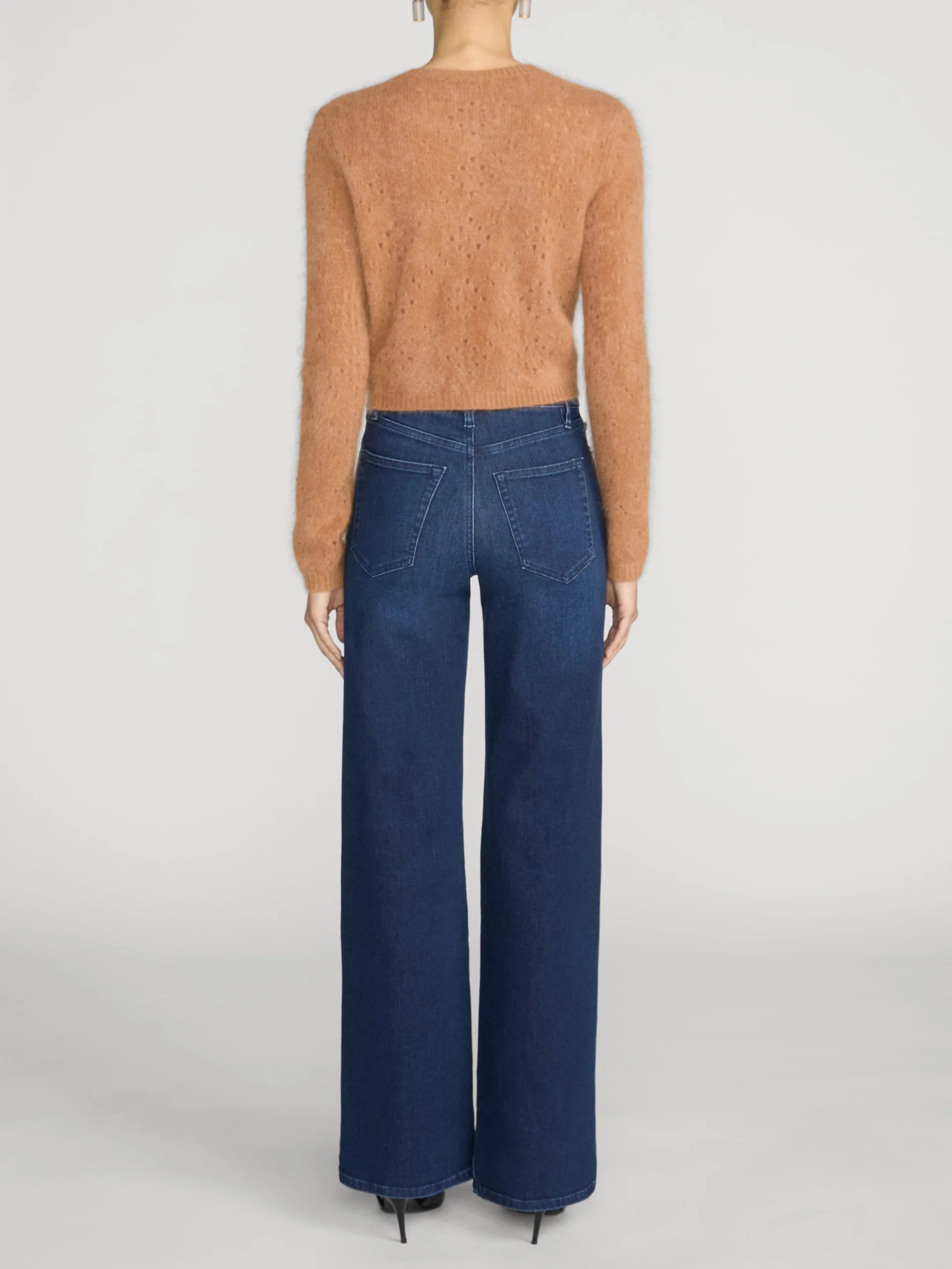 A model shown from the back wearing a fuzzy cropped long-sleeve sweater in a warm camel tan and dark blue wide-leg jeans, set against a plain light gray studio background.
