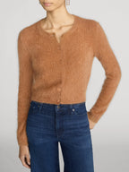 A model wearing a fuzzy, cropped long-sleeve cardigan in a warm camel brown color, paired with dark blue jeans, against a light gray studio background.