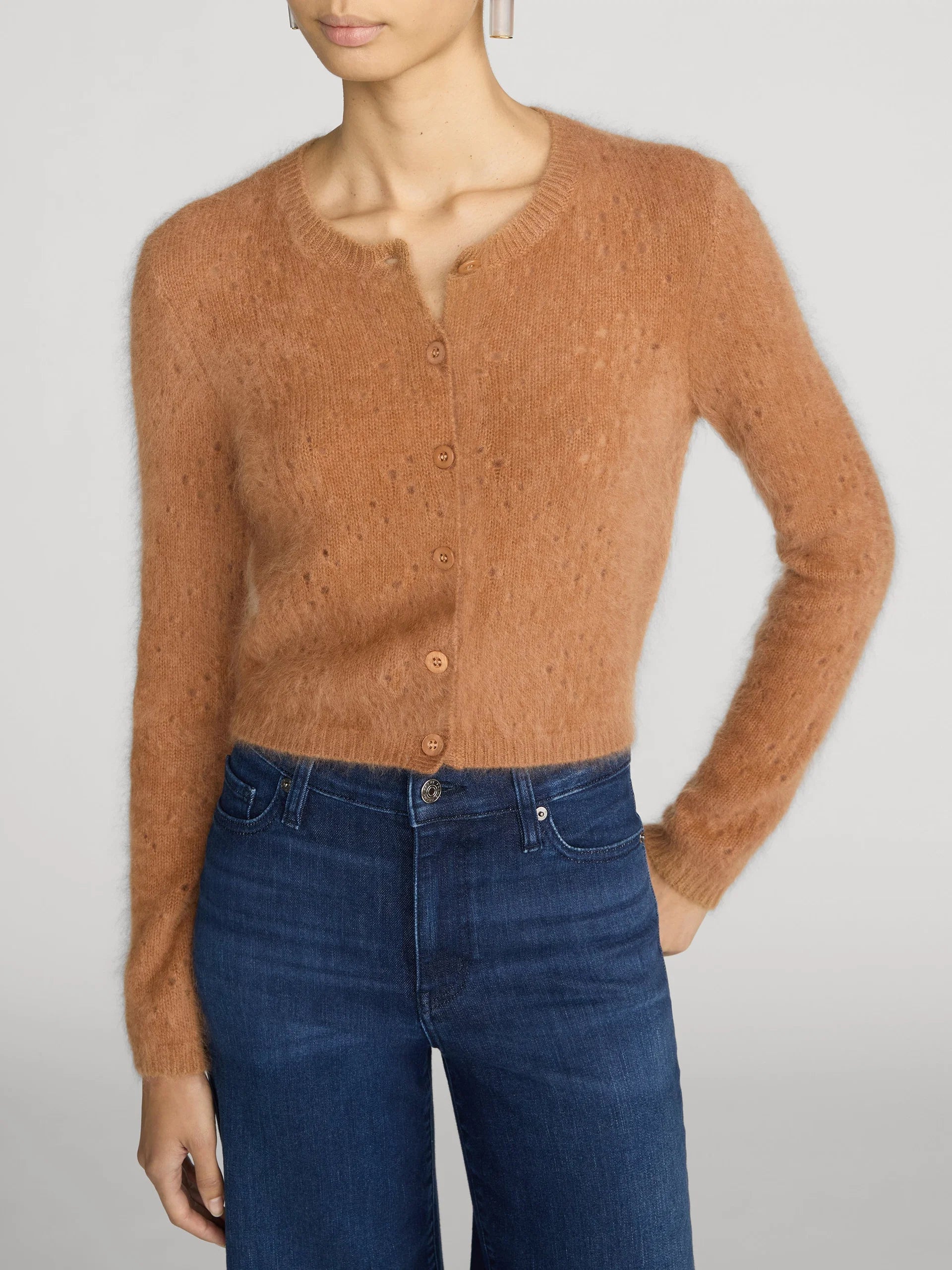 A model wearing a fuzzy, cropped long-sleeve cardigan in a warm camel brown color, paired with dark blue jeans, against a light gray studio background.