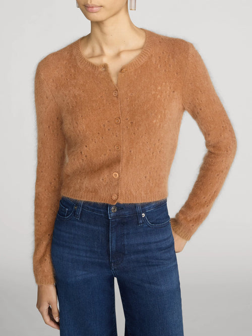 A model wearing a fuzzy, cropped long-sleeve cardigan in a warm camel brown color, paired with dark blue jeans, against a light gray studio background.