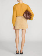 A model shown from the back wearing a mustard yellow long-sleeve blouse, a beige mini skirt, and black heels, while carrying a brown leather clutch bag against a light gray studio background.
