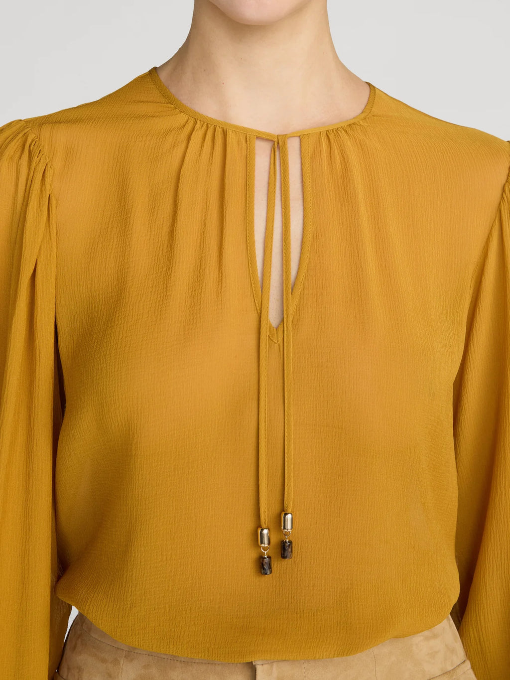 A close-up of a mustard yellow blouse with long sleeves, a round neckline, and a front keyhole cutout with tie details and small metallic bead accents.