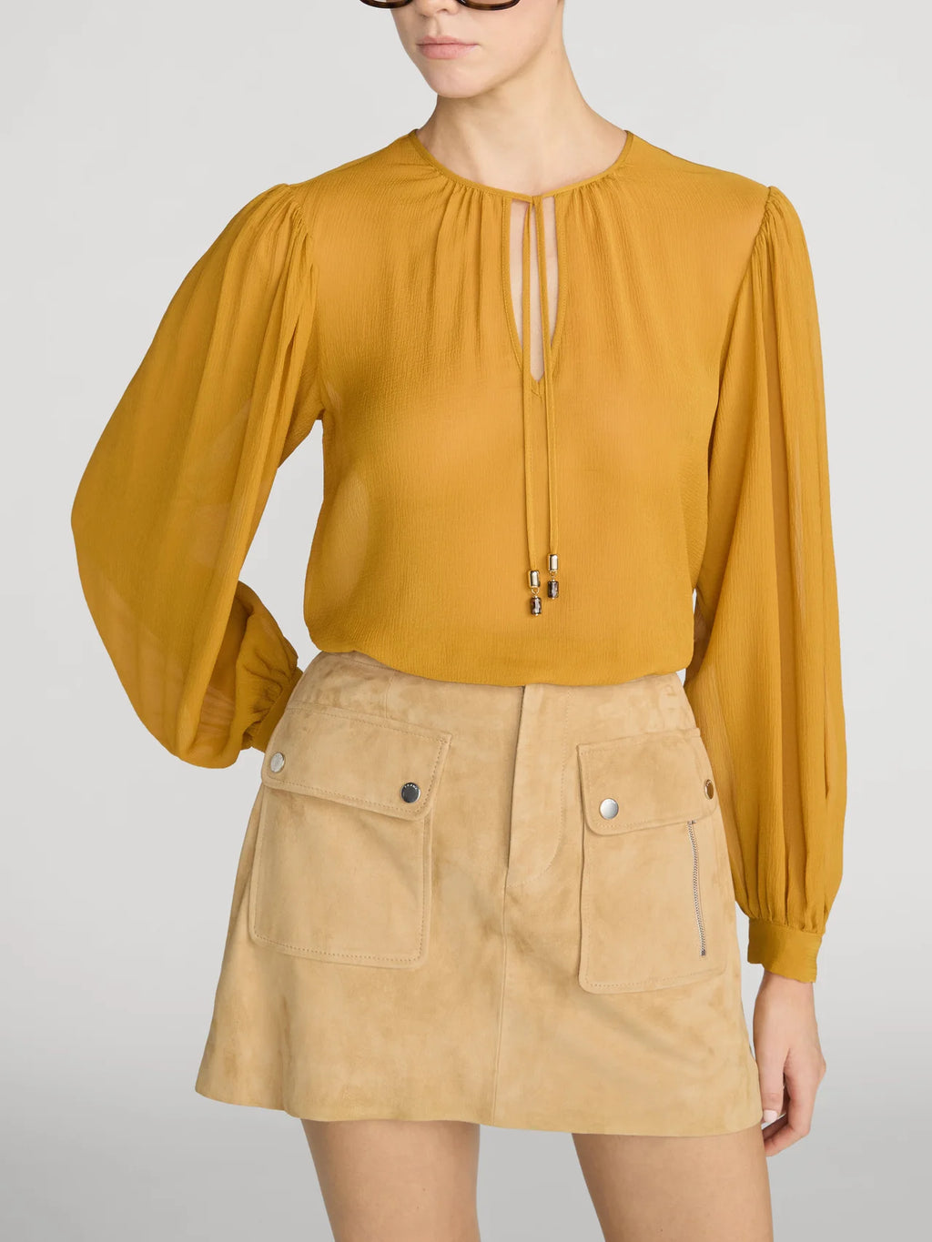 A model wearing a mustard yellow long-sleeve blouse with a keyhole front and a tan mini skirt with front flap pockets, set against a plain light gray studio background.