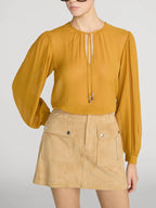 A model wearing a mustard yellow long-sleeve blouse with a keyhole front and a tan mini skirt with front flap pockets, set against a plain light gray studio background.