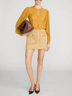 A model wearing a mustard yellow long-sleeve blouse with a tie-neck detail, a beige mini skirt with front flap pockets, and holding a brown leather clutch against a light gray studio background.