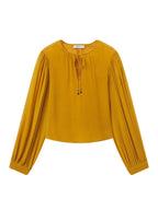A mustard yellow long-sleeve blouse with a round neckline, front tie detail, and gathered sleeves, shown flat on a light gray background.