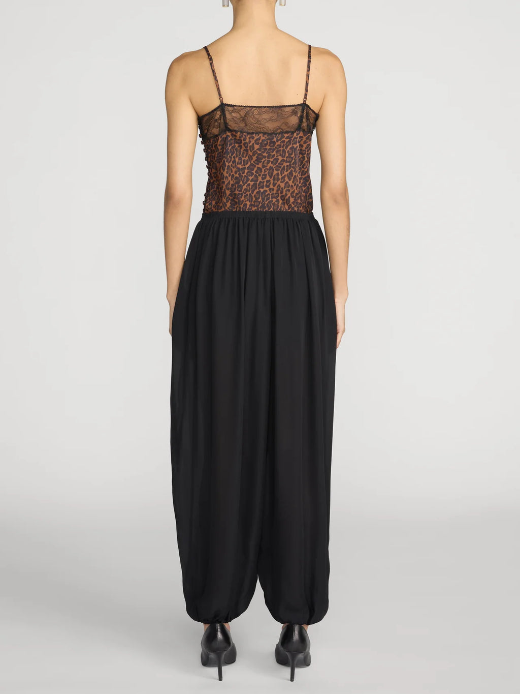 A model shown from the back wearing a black harem-style jumpsuit with a sheer leopard-print bodice and thin spaghetti straps, paired with black pointed-toe heels, against a light gray studio background.