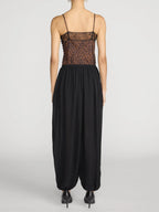 A model shown from the back wearing a black harem-style jumpsuit with a sheer leopard-print bodice and thin spaghetti straps, paired with black pointed-toe heels, against a light gray studio background.