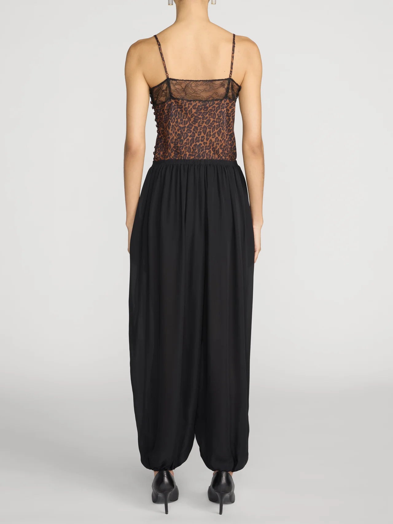 A model shown from the back wearing a black harem-style jumpsuit with a sheer leopard-print bodice and thin spaghetti straps, paired with black pointed-toe heels, against a light gray studio background.