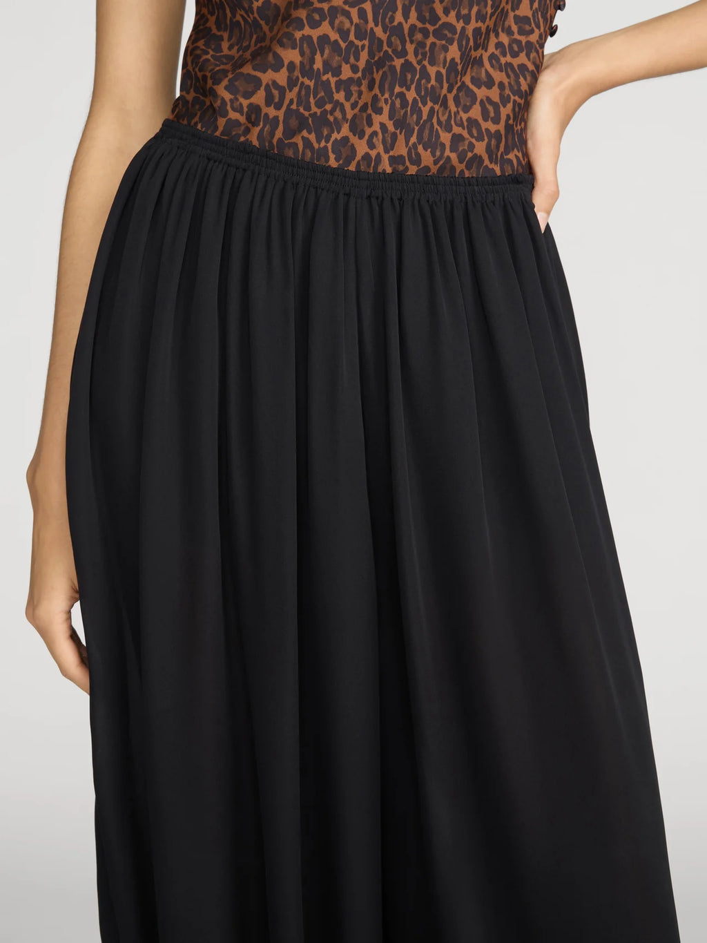 A close-up of a model wearing a black pleated skirt with a leopard-print top, shown from the waist down against a light gray background.