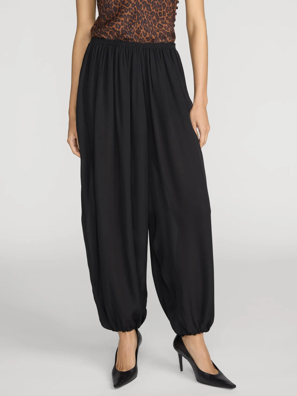 A model wearing black harem-style pants with an elastic waist and gathered cuffs, paired with a leopard-print top and black heels, against a light gray studio background.