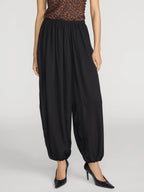 A model wearing black harem-style pants with an elastic waist and gathered cuffs, paired with a leopard-print top and black heels, against a light gray studio background.